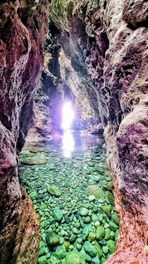 Interior de la Cueva de los Colores con agua cristalina, rocas volcánicas y entrada de luz natural al fondo.